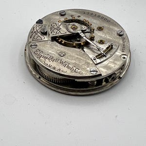 May include: Close-up of an antique Elgin Natl. Watch Co. pocket watch movement. The silver-toned mechanism features intricate gears, jewels, and the text "15 Jewels." The watch movement is marked "USA" and has the serial number 13418900.