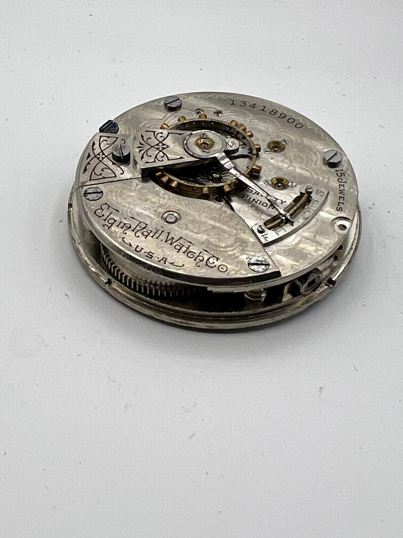 May include: Close-up of an antique Elgin Natl. Watch Co. pocket watch movement. The silver-toned mechanism features intricate gears, jewels, and the text "15 Jewels." The watch movement is marked "USA" and has the serial number 13418900.