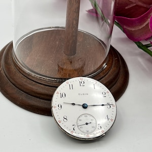 May include: An antique Elgin pocket watch face with a white dial, black hands, and a subdial. The watch face is set against a wooden base, with a glass cloche and a wooden stem. The watch face is approximately 2 inches in diameter.