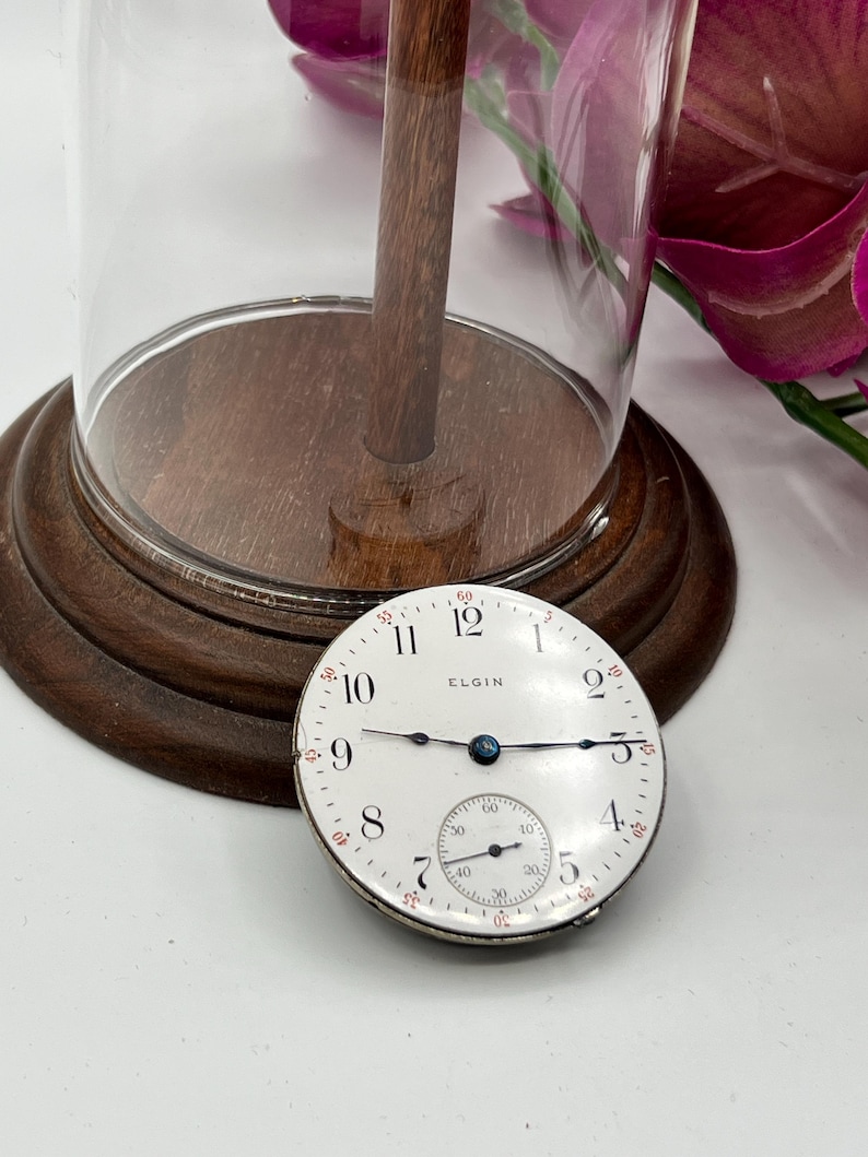 May include: An antique Elgin pocket watch face with a white dial, black hands, and a subdial. The watch face is set against a wooden base, with a glass cloche and a wooden stem. The watch face is approximately 2 inches in diameter.