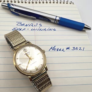 May include: A gold-tone Benrus self-winding wristwatch with a metallic expansion band, featuring a round white dial with a date window at the twelve o'clock position. The watch is placed on lined paper with handwritten text reading Benrus Self-Winding and Model # 3021.