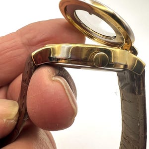 May include: A gold-toned wristwatch with a brown leather band. The watch face is covered by a hinged, clear cover. The watch has a round face and a small button on the side. The band is a dark brown color.
