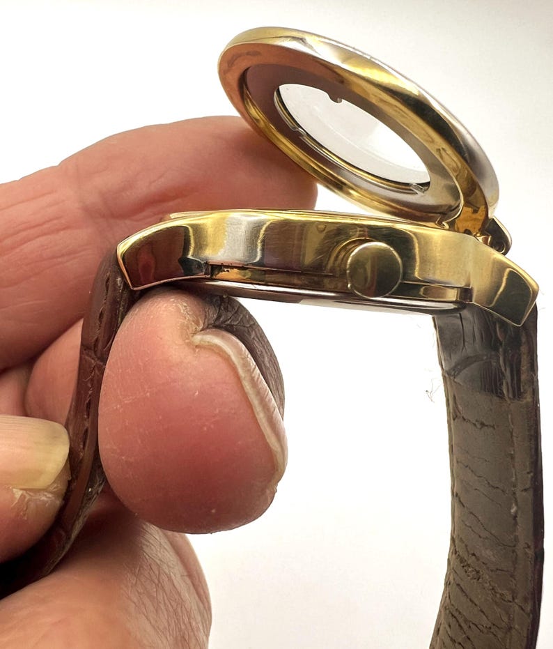 May include: A gold-toned wristwatch with a brown leather band. The watch face is covered by a hinged, clear cover. The watch has a round face and a small button on the side. The band is a dark brown color.