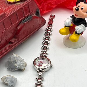 Op de afbeelding: Een zilverkleurig polshorloge met een Mickey Mouse-ontwerp op de wijzerplaat en een decoratieve schakelband. De band heeft kleine, ronde elementen. Een rode speelgoedbrandweerwagen en een Mickey Mouse-beeldje zijn ook zichtbaar.