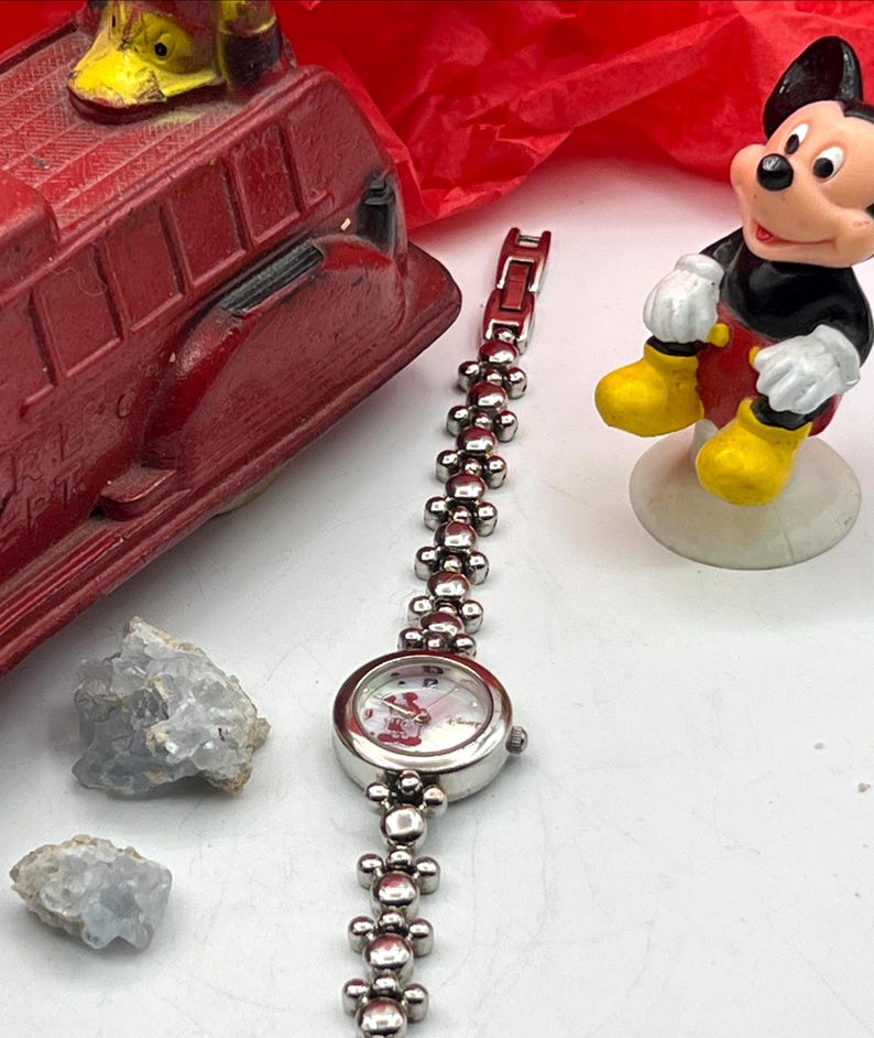 Op de afbeelding: Een zilverkleurig polshorloge met een Mickey Mouse-ontwerp op de wijzerplaat en een decoratieve schakelband. De band heeft kleine, ronde elementen. Een rode speelgoedbrandweerwagen en een Mickey Mouse-beeldje zijn ook zichtbaar.