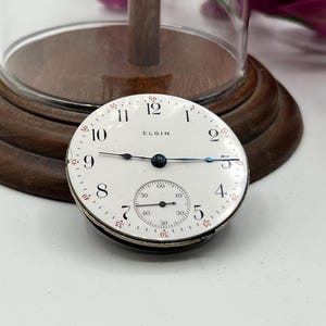 May include: An antique Elgin pocket watch face with a white dial, black numerals, and blue hands. The watch face includes a smaller subdial and the word "ELGIN". The watch face is set against a wooden base.