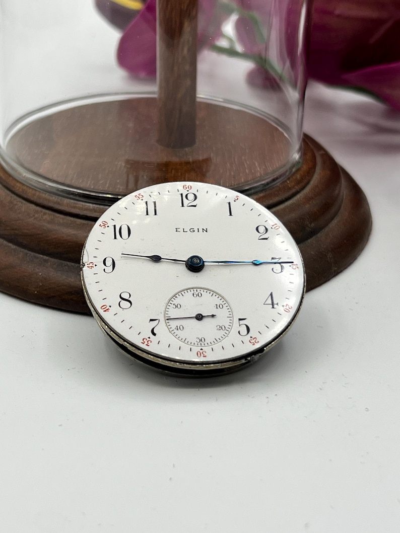 May include: An antique Elgin pocket watch face with a white dial, black numerals, and blue hands. The watch face includes a smaller subdial and the word "ELGIN". The watch face is set against a wooden base.
