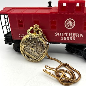 Puede incluir: Un reloj de bolsillo dorado con un grabado de tren, cadena y cierre. El reloj está delante de un vagón de tren de juguete rojo con el texto "SOUTHERN X9066". El reloj tiene un diseño ornamentado.