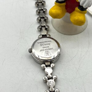 Op de afbeelding: Zilverkleurig Disney-horloge met een Mickey Mouse-thema band. De wijzerplaat is rond en de band heeft Mickey Mouse-hoofdvormen. De achterkant van het horloge is gemarkeerd met "Stainless Steel Back", "Disney" en "Japan Movement".