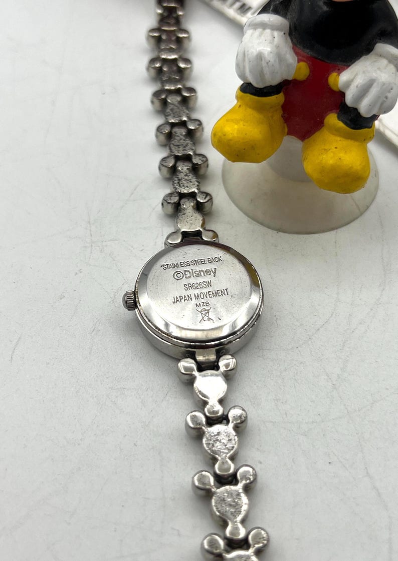 Op de afbeelding: Zilverkleurig Disney-horloge met een Mickey Mouse-thema band. De wijzerplaat is rond en de band heeft Mickey Mouse-hoofdvormen. De achterkant van het horloge is gemarkeerd met "Stainless Steel Back", "Disney" en "Japan Movement".
