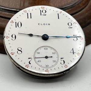 May include: A vintage Elgin pocket watch face with a white dial, black numerals, and a smaller seconds dial. The watch face has black and blue hands and the brand name "ELGIN" is printed on the dial.