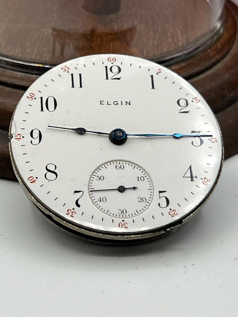 May include: A vintage Elgin pocket watch face with a white dial, black numerals, and a smaller seconds dial. The watch face has black and blue hands and the brand name "ELGIN" is printed on the dial.