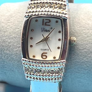 Può includere: Orologio al quarzo Vivani color argento con quadrante rettangolare. L'orologio ha un quadrante bianco con numeri neri alle 12 e alle 6 e lancette dorate. Il cinturino argentato ha strass decorativi.