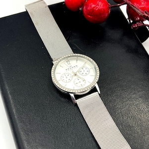 May include: A silver Skagen watch with a white face and a mesh band. The watch face has three sub-dials and is surrounded by a ring of small, clear stones. The watch is displayed on a black surface.
