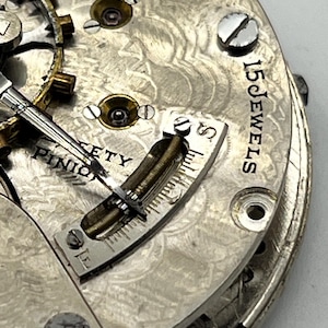 May include: Close-up of a vintage watch movement. The silver-toned mechanism features intricate details, including gears, screws, and a balance wheel. The text "15 JEWELS" is visible, along with other markings. The watch has a detailed, engraved pattern.