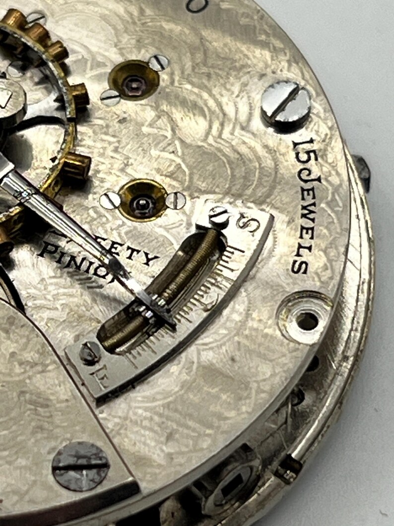 May include: Close-up of a vintage watch movement. The silver-toned mechanism features intricate details, including gears, screws, and a balance wheel. The text "15 JEWELS" is visible, along with other markings. The watch has a detailed, engraved pattern.