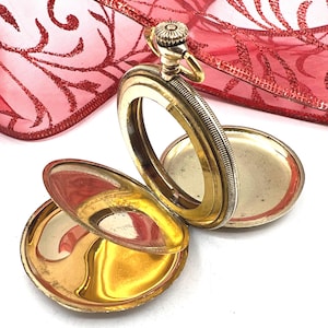 May include: An open gold-tone pocket watch case with a hinged cover and a circular frame. The metal has a polished finish with a textured edge on the winding crown and the outer rim. The interior of the case is smooth and reflective.