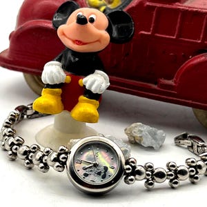 Op de afbeelding: Een zilverkleurig Mickey Mouse-horloge met een decoratieve band en een parelmoeren wijzerplaat. Een Mickey Mouse-beeldje en een rode speelgoedbrandweerwagen staan op de achtergrond. Het horloge is gemarkeerd "China".