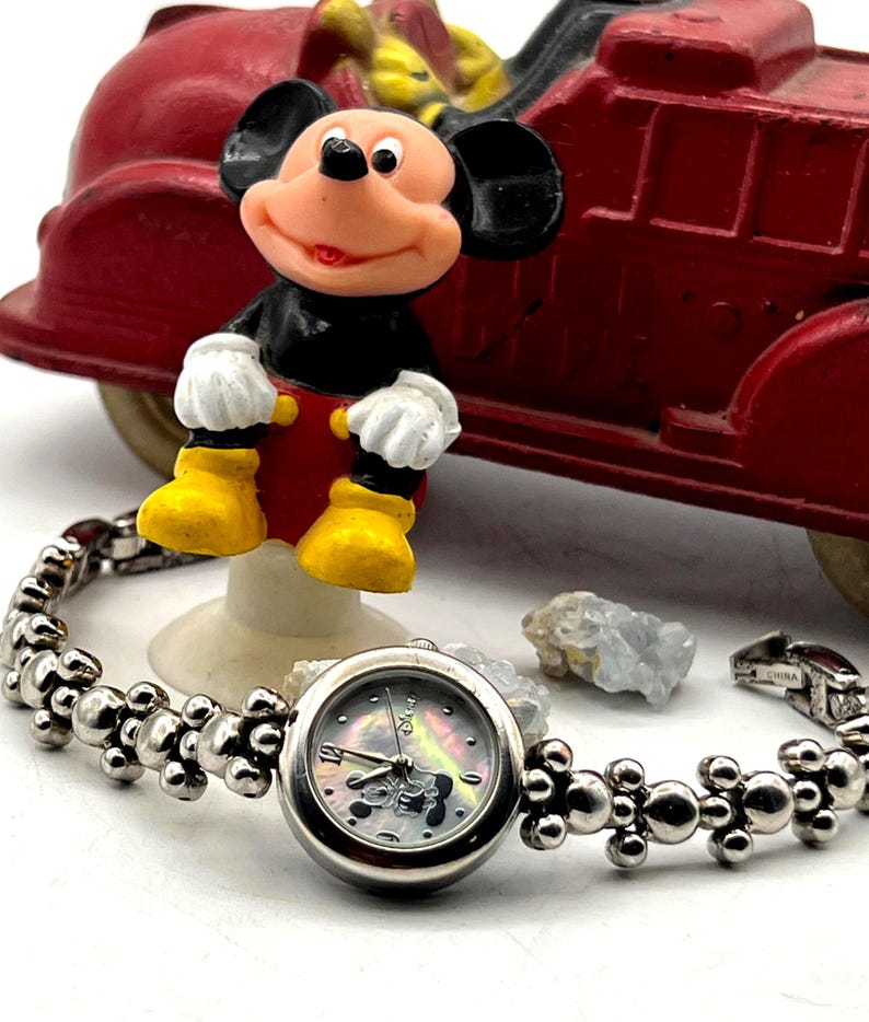 Op de afbeelding: Een zilverkleurig Mickey Mouse-horloge met een decoratieve band en een parelmoeren wijzerplaat. Een Mickey Mouse-beeldje en een rode speelgoedbrandweerwagen staan op de achtergrond. Het horloge is gemarkeerd "China".