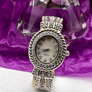 Könnte beinhalten: Eine verzierte Quarzuhr mit einem silber- und goldfarbenen Metallarmband und Zifferblatt. Das ovale Zifferblatt hat einen weißen Hintergrund mit schwarzen Ziffern und Zeigern. Das Armband hat dekorative Schnörkel und kleine klare Kristalle.