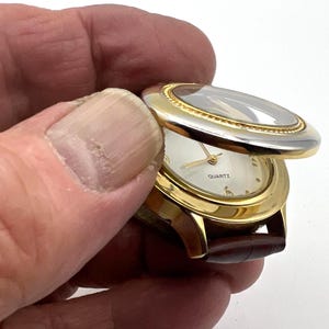 May include: A gold-toned quartz watch with a brown leather band. The watch face is white with gold hands and hour markers. The watch has a hinged cover with a clear crystal. The word "QUARTZ" is printed on the watch face.