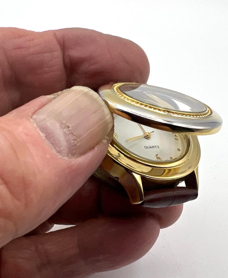 May include: A gold-toned quartz watch with a brown leather band. The watch face is white with gold hands and hour markers. The watch has a hinged cover with a clear crystal. The word "QUARTZ" is printed on the watch face.