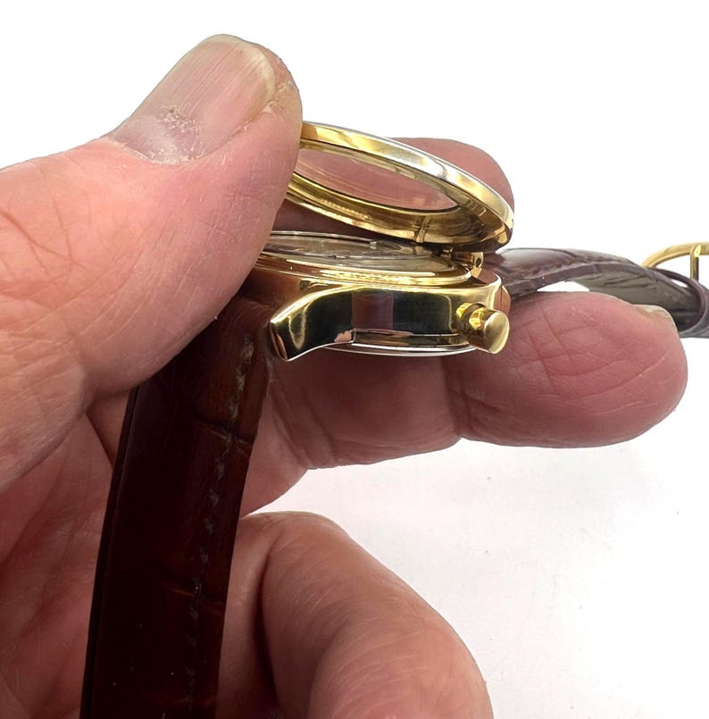 May include: A gold-toned wristwatch with a brown leather band. The watch face is visible through a clear cover, and the band is secured with a gold-colored buckle. The watch is held open, revealing the watch face.