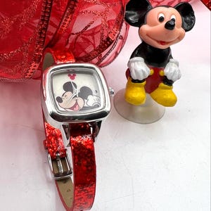 Puede incluir: Un reloj cuadrado plateado con una correa roja brillante, con un diseño de Mickey Mouse en la esfera. Una pequeña figura de Mickey Mouse está cerca. La correa del reloj tiene un cierre de hebilla.