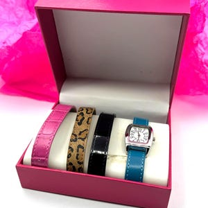 May include: A watch set in a pink box. The set includes a silver watch with a teal band, and three additional bands in pink, leopard print, and black. The watch face is white with black numbers.