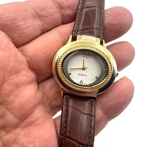 May include: Gold-tone quartz watch with a white face and brown leather band. The watch face features gold accents, black numerals, and gold hands. The band has a crocodile texture.