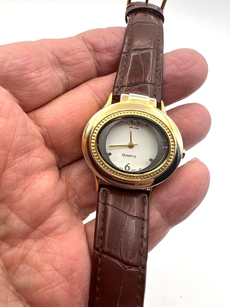 May include: Gold-tone quartz watch with a white face and brown leather band. The watch face features gold accents, black numerals, and gold hands. The band has a crocodile texture.