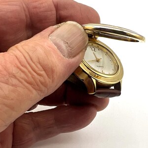 May include: A gold-toned quartz watch with a brown leather band. The watch face is oval with gold hands and a white background. The watch has a hinged cover that is open, revealing the watch face. The watch is held in a hand.