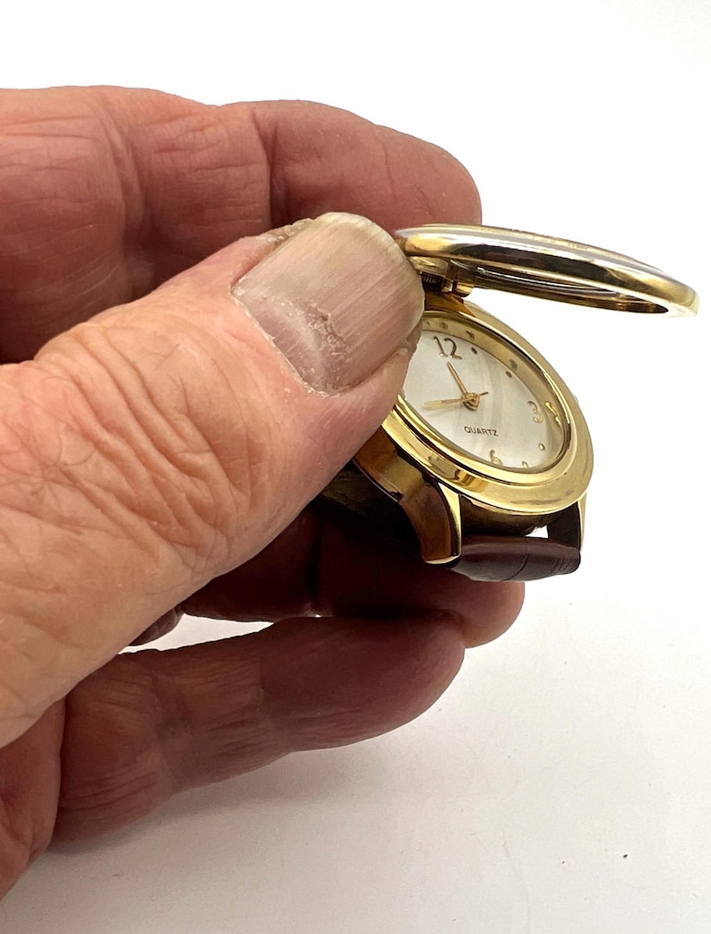May include: A gold-toned quartz watch with a brown leather band. The watch face is oval with gold hands and a white background. The watch has a hinged cover that is open, revealing the watch face. The watch is held in a hand.
