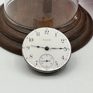 May include: A vintage Elgin pocket watch face with a white dial, black hands, and black numerals. The watch face has a subdial and the word "ELGIN" is printed on the face. The watch face is displayed on a wooden base.