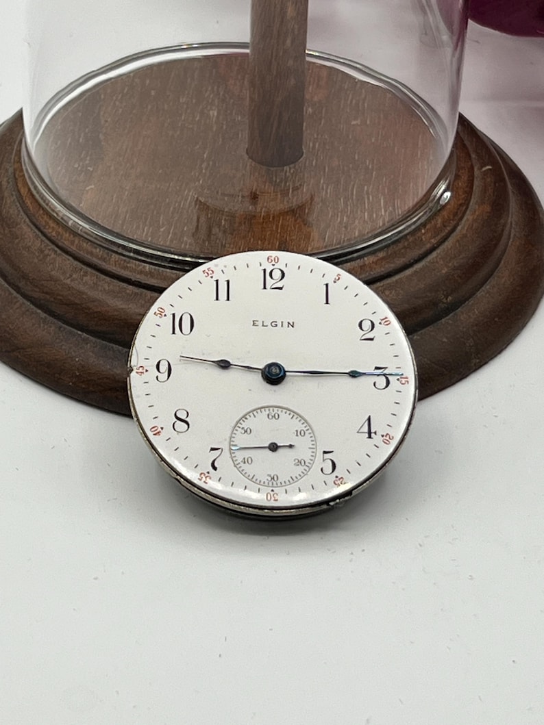 May include: A vintage Elgin pocket watch face with a white dial, black hands, and black numerals. The watch face has a subdial and the word "ELGIN" is printed on the face. The watch face is displayed on a wooden base.