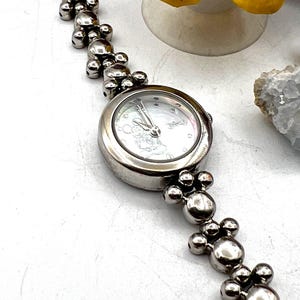 Op de afbeelding: Zilverkleurig polshorloge met een ronde wijzerplaat en een Mickey Mouse-ontwerp. De horlogeband heeft een reeks verbonden, bolvormige kralen. Het horloge wordt op een witte ondergrond getoond.