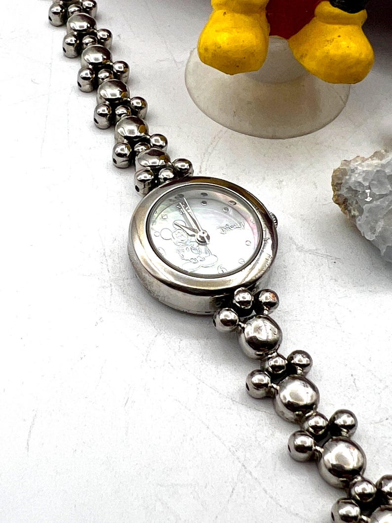Op de afbeelding: Zilverkleurig polshorloge met een ronde wijzerplaat en een Mickey Mouse-ontwerp. De horlogeband heeft een reeks verbonden, bolvormige kralen. Het horloge wordt op een witte ondergrond getoond.