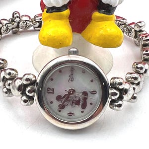 Op de afbeelding: Een zilverkleurig Disney-horloge met een ronde wijzerplaat en een armband. De wijzerplaat heeft een Mickey Mouse-ontwerp en het Disney-logo. De band heeft een uniek, gestructureerd ontwerp. Het horloge is een charmant accessoire.