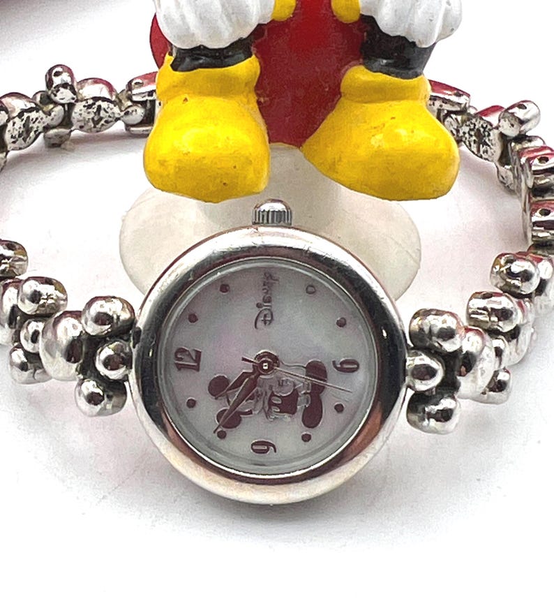 Op de afbeelding: Een zilverkleurig Disney-horloge met een ronde wijzerplaat en een armband. De wijzerplaat heeft een Mickey Mouse-ontwerp en het Disney-logo. De band heeft een uniek, gestructureerd ontwerp. Het horloge is een charmant accessoire.