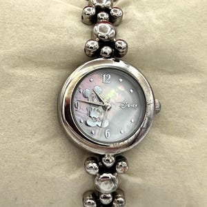 Op de afbeelding: Zilverkleurig Disney-horloge met een Mickey Mouse-ontwerp op de wijzerplaat. Het horloge heeft een ronde wijzerplaat met een parelmoereffect en een band in armbandstijl. De band heeft ronde, decoratieve elementen.