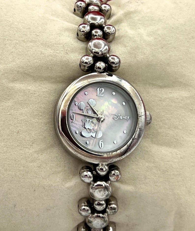 Op de afbeelding: Zilverkleurig Disney-horloge met een Mickey Mouse-ontwerp op de wijzerplaat. Het horloge heeft een ronde wijzerplaat met een parelmoereffect en een band in armbandstijl. De band heeft ronde, decoratieve elementen.