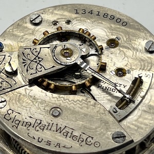 May include: Close-up of an antique Elgin Rail Watch Co. pocket watch movement. The silver-toned mechanism features intricate gears, screws, and the text "13418900". The watch is marked "U.S.A."