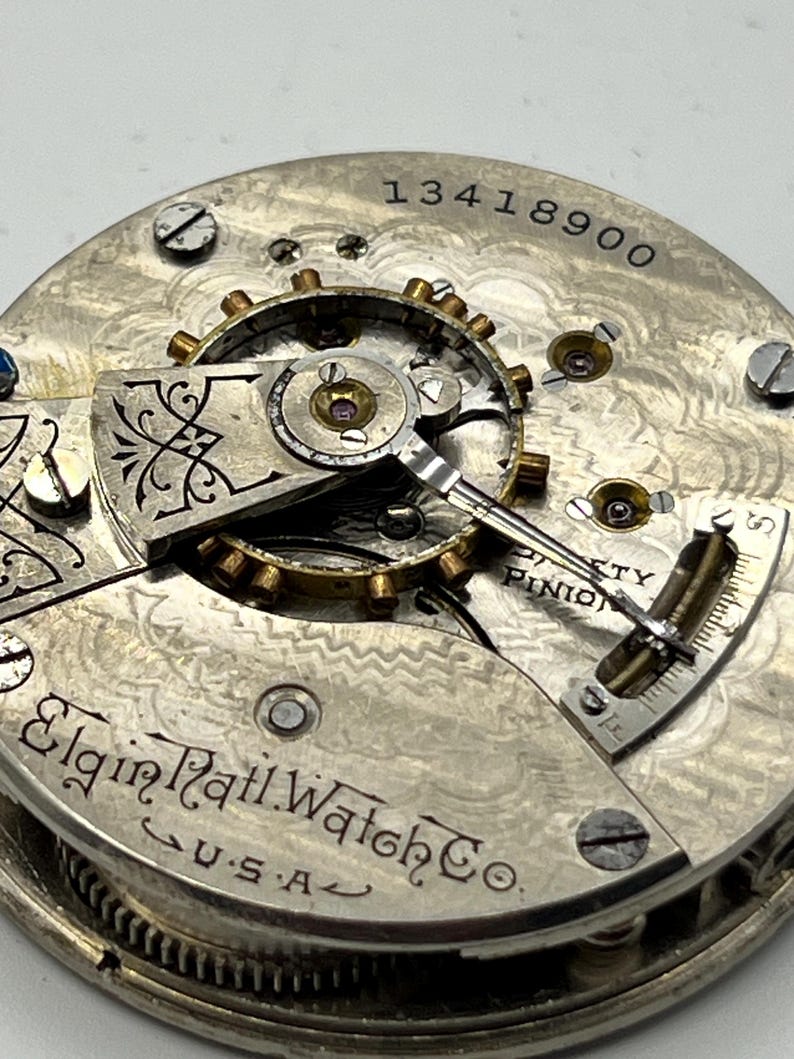 May include: Close-up of an antique Elgin Rail Watch Co. pocket watch movement. The silver-toned mechanism features intricate gears, screws, and the text "13418900". The watch is marked "U.S.A."