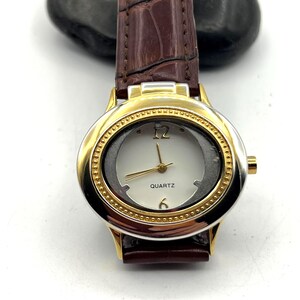 May include: A gold-toned quartz watch with a brown leather band. The watch face is white with gold hands and the numbers 12 and 6. The band has a crocodile skin texture.