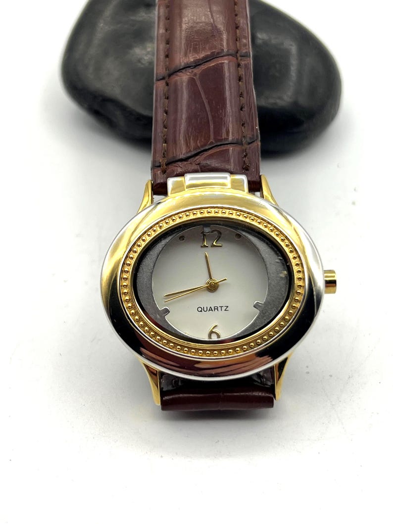 May include: A gold-toned quartz watch with a brown leather band. The watch face is white with gold hands and the numbers 12 and 6. The band has a crocodile skin texture.