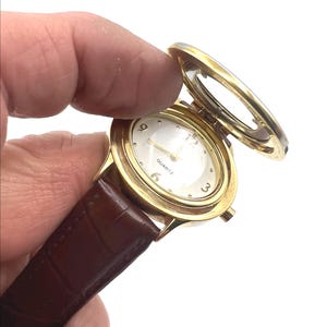 May include: A gold-toned quartz watch with a brown leather band. The watch face is white with gold hands and numerals. The watch has a hinged cover that is open, revealing the watch face. The watch is held by a hand.