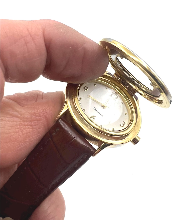 May include: A gold-toned quartz watch with a brown leather band. The watch face is white with gold hands and numerals. The watch has a hinged cover that is open, revealing the watch face. The watch is held by a hand.