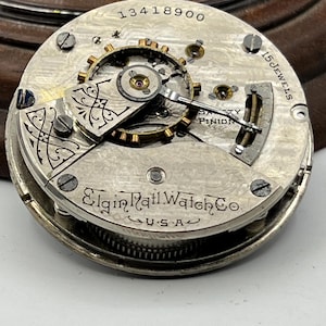 May include: Close-up of a vintage Elgin Rail Watch Co. movement. The silver-toned mechanism features intricate gears, screws, and the text "15 Jewels" and "U.S.A.". The serial number "13418900" is visible.