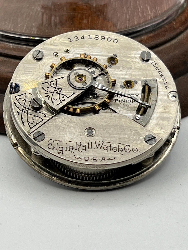 May include: Close-up of a vintage Elgin Rail Watch Co. movement. The silver-toned mechanism features intricate gears, screws, and the text "15 Jewels" and "U.S.A.". The serial number "13418900" is visible.
