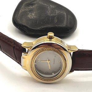 May include: A gold-toned quartz watch with a silver face and gold hands. The watch has a brown leather band. A dark gray stone is in the background.